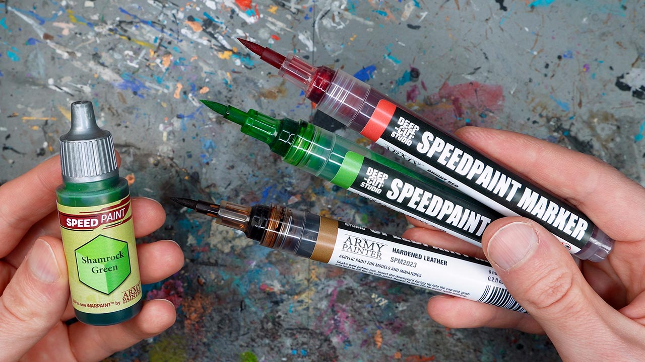 Left hand holding a bottle of Shamrock Green Speedpaint from The Army Painter, right hand holding three Speedpaint markers, grey speckled background