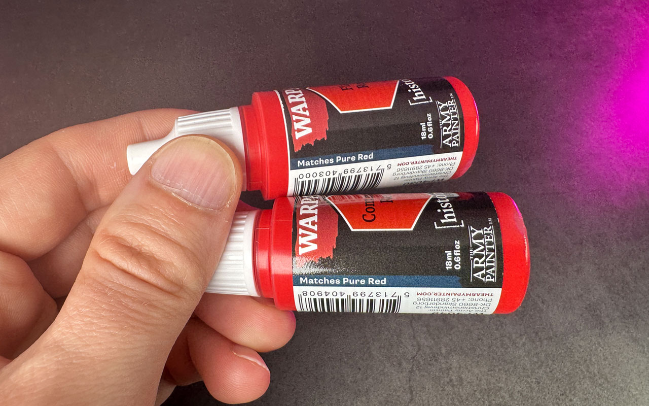 A hand holding bottles of Warpaints Fanatic Ccommand Red and Flag red paint bottles, comparing the labels