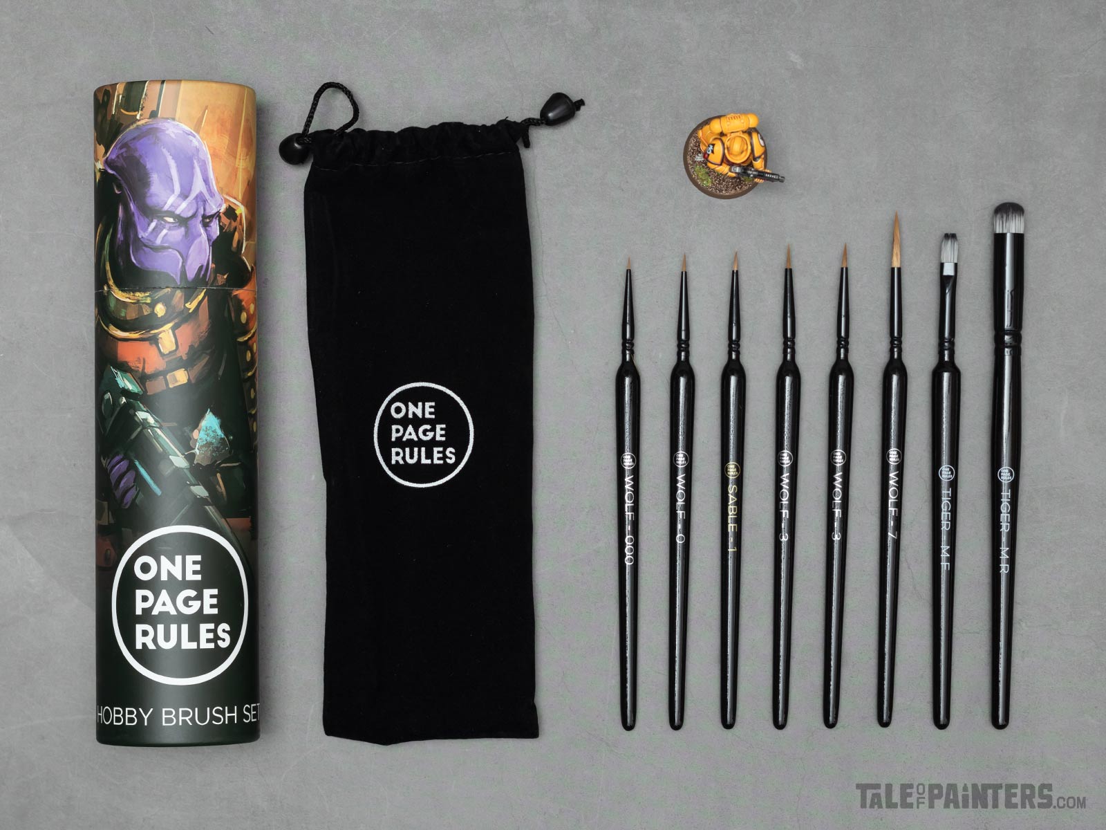The contents of Chronicle's One Page Rules brush set laid out flat on a concrete background: box, black velvet pouch, and seven brushes from small to large, with a Primaris Space Marine model for size comparison