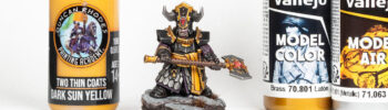 A Helsmith of Hashut Infernal Cohort model painted in the colours of Muspelzharr, surrounded by Vallejo and Two Thin Coats paint bottles, arranged for a painting tutorial