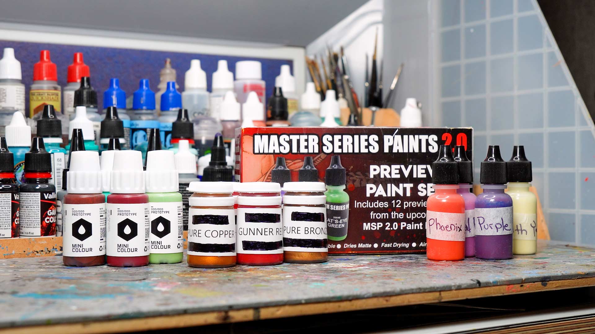 A workbench filled with various prototype miniature paints, from left to right: Mini Colour, P3 wave 2, Reaper Master Series 2.0 bottles