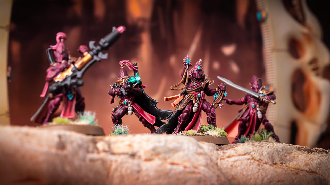 Cinematic model shot of four Aeldari Corsairs in burgundy armour, inc. a Soul Weaver, standing atop a sandy cliff, warm orange background with lots of Aeldari architecture